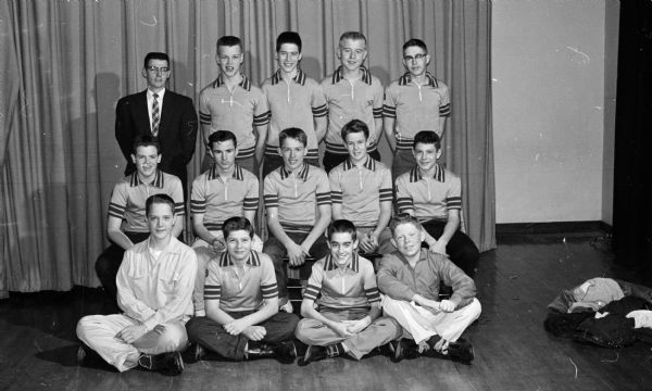 Thirteen teenage boys wear matching shirts with striped collars, sleeve bands, and waist bands to pose in three rows with their coach. The four boys standing in back are all taller than their coach. The Frank Allis grade school basketball team on Buckeye Road won 13 straight victories in the Suburban Elementary League. Shown (L-R) front row, are: Doug McLeisel (manager), Brian Schroeder, Gary Johnson, and Darrell Vogt (manager). Shown (L-R) second row, are: Bob Branch, Eddie Francis, Tom McKay, Mike Roy, and Dennis Eley. Shown (L-R) in the back row, are: Coach Irv Kennedy (wearing a suit coat and neck tie), Tim Walterscheidt, Rockey Meives, Dave Tipple, and Greg Quinn.