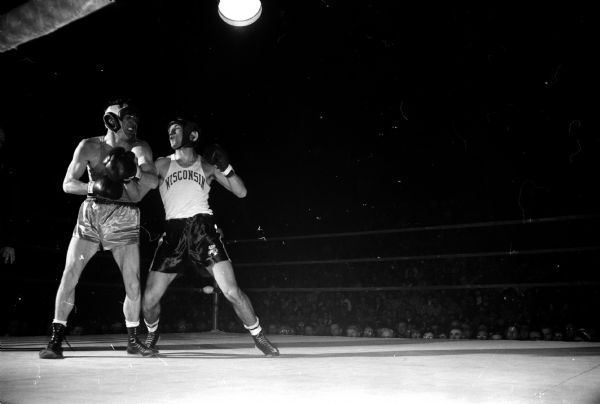Boxer Charlie Mohr blocks a punch with his right arm while winding up to deliver a left jab to his opponent. Both boxers wear leather helmets. The original caption states: "Wisconsin's Charlie Mohr (white shirt) has hurt Sacramento State's Dave Smith with a right to the body and is about to land a left to the head. Mohr won all the way in this fight at 165 pounds when the Badgers took a 6-2 team victory."