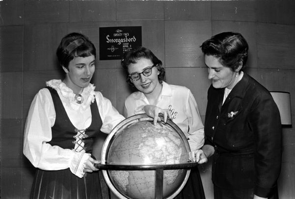 Three women, one wearing Scandinavian folk clothing, are examining a globe while making plans for the U.W. YWCA's benefit smorgasbord. The event will help finance students attending conferences and support the YWCA's work with international students. The three women are Sinikka Pesola, a student from Finland, and Darlene Conner, general chairman, and Martha Newell, executive secretary of the University YWCA.