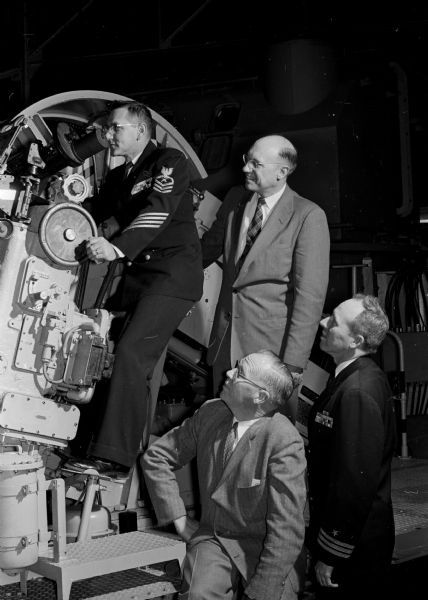 Madison men who play naval officers in the play "South Pacific" learn about naval weapons by visiting the Naval Reserve Officers Training corps (NROTC) headquarters at the University of Wisconsin. Shown are Chief Gunner's Mate Emil J. Szpila, an instructor, demonstrating the operation of a dual purpose anti-aircraft gun to actor Jerry Dame, standing behind Szpila, and actor Victor Johnson, sitting to the right of Szpila, and Cmdr. John E. O'Neill, executive officer of the NROTC.