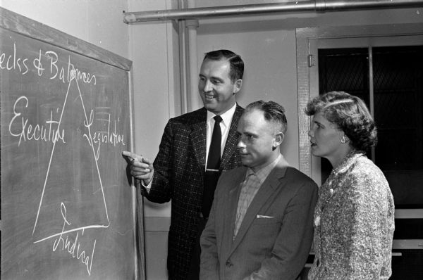 Richard D. Carleton, left, instructor from the Madison Vocational school, stands at a blackboard with two of his students, Frank Vanhontte, from Belgium, and Irmgard Hall, from Germany. The students recently completed an American citizenship course offered by the Truax Field Education center.