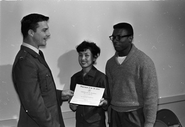 Lt. Charles Grimminger Jr., Truax education officer, left, hands a Certificate of Training to Mrs. Midori Fugate of Japan and Romeo Downer from British West Indies. They have completed an American citizenship course offered by Truax Field Education center.