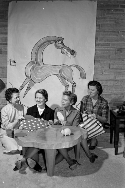 Working on plans for the annual dinner meeting of the Wisconsin Alumni Club of Madison are Anya Castle, Dorothy Curreri, Mary Mohs, and Jean Johnson. The speaker, Donald Slichter, will speak on "Piggy Banks and Gift Horses." An Aaron Bohrod depiction of a horse with a gift in its mouth is on the wall in the background.