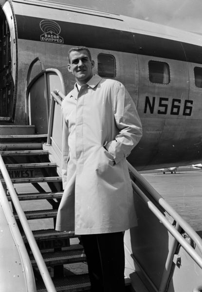 A man (Henry Derleth) wears an overcoat while posed on the middle of the boarding stairway attached to a partially visible passenger airliner. The original caption states, "Henry Derleth, star end on the University of Wisconsin's 1959 Big 10 football champion teams, boards a plane at Madison Municipal airport en route to New York and London. Derleth is one of six outstanding athletes in the nation selected to serve as counselors and escorts for 72 newspaperboys who won trips to London and Paris in the 1960 'Young Columbus IV' contest sponsored by Parade magazine and their local newspapers."