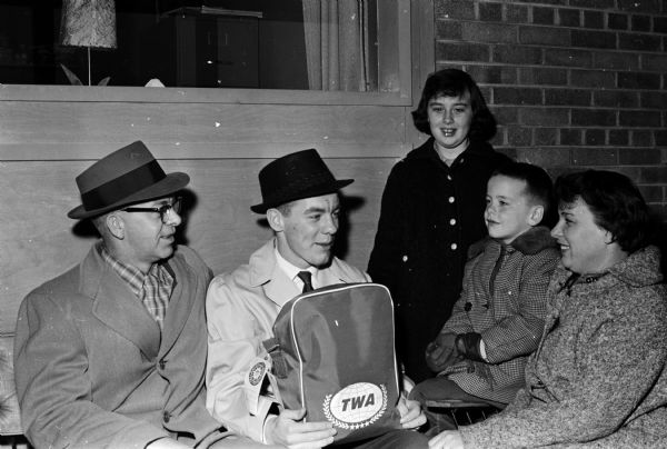 Contest winner George Byrne, surrounded by his family, wears an overcoat, white shirt, tie and fedora (hat) while holding a TWA travel bag. The original caption states: "a happy 'Young Columbus IV' took off from Madison's Truax Field bound for the Old World. George Byrne, 15, Brooklyn (Wisconsin), won the  eight day flying trip to Paris and London for selling 201 newspaper subscriptions in his home town. George is a newspaper carrier for The Wisconsin State Journal. George is shown at the airport with his family, from left to right: Mr. Cletus Bryne, his father, George, his 10-year-old sister, Patricia, his brother, Michael, 5, and Mrs. Byrne. The contest which George won is sponsored by Madison Newspapers,  Parade magazine and Trans World Airlines (TWA)."