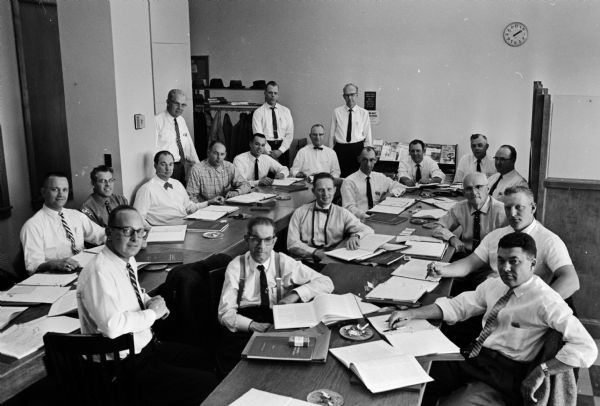 A group of twenty men sit on either side of long tables while taking a three month training course in accident and health insurance underwriting.