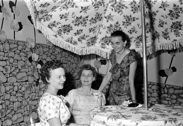 Three committee members for the East Side Women's Club's spring banquet chat beneath a large umbrella in a garden-like setting. They include, from left: Kathleen Olson, Eleanor Esser, and Mrs. Donald Green.