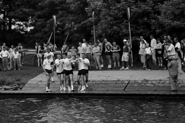 View across water towards the victorious and still undefeated California Golden Bears shoulder their coxswain Arlen Lackey, and are heading down a boat ramp. Their crew finished first and was clocked in 9 minutes, five-tenths of a second over a nine and three-quarters course on Lake Monona.
