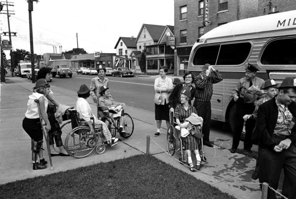 Madison firemen from Station No. 3 at 1217 Williamson Street, along with their wives, took 32 handicapped children to the Braves-Los Angeles baseball game in Milwaukee. The firemen's wives are pushing the wheelchairs toward the bus. In the background are houses and businesses in the 1200 block of Williamson Street, including 1206 Williamson General Sales & Supply Co., 1210 Williamson Venetian Cafe, 1216 Williamson Lee Cleaners.
