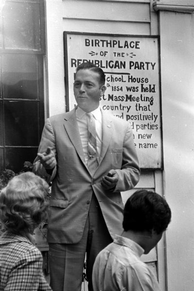 Ivan Kindschi, Dane County dairy farmer and candidate for state treasurer, speaks from the steps of the Little White Schoolhouse as the GOP candidates begin a five day team campaign. A sign behind him is titled "Birthplace of the Republican Party".