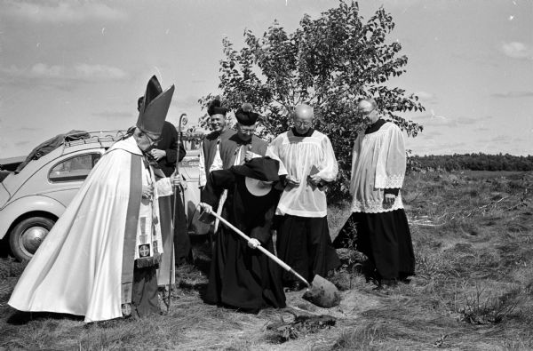 Mother Monica, superior of the convent and academy, turns over the first spadeful of ground during the groundbreaking ceremonies for a new motherhouse convent for the Academy of St. Benedict at Fox Bluff. Others taking part in the ceremony are, left to right: Bishop William P. O'Connor of the Madison Catholic diocese; Rev. Wilfred Tunink, St. Louis, Missouri (partially hidden); Msgr. Edward B. Auchter, St. Mary of the Lake Church, Westport; Msgr. Ferdinand A. Mack, St. Bernard's Church, Middleton; Rev. Justin Sion, academy chaplain; and Rev. Joseph L. Brechtl, St. Andrew's Catholic Church, Verona.