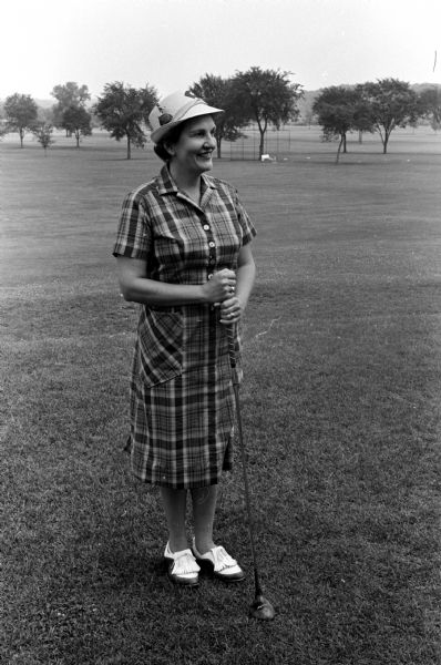 Margorie Hagan, general chairman of the women's organization of the Maple Bluff Country Club, wearing an attractive plaid golf dress. She also models a decorated hat and is holding a golf club.