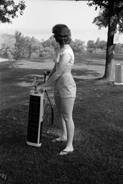 An unnamed woman is holding a golf bag while modeling what a female golfer should NOT wear on the golf course: a halter top, tight shorts, and sandals.
