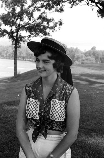 Joan Schlicht modeling a straw hat with a dangling hat band and a patterned blouse.