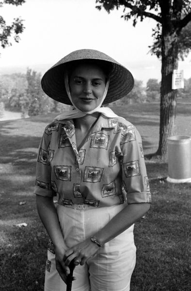 Ruth Buslee modeling a cone shaped straw hat held on her head by a fabric chin tie, along with a printed blouse and a necklace.