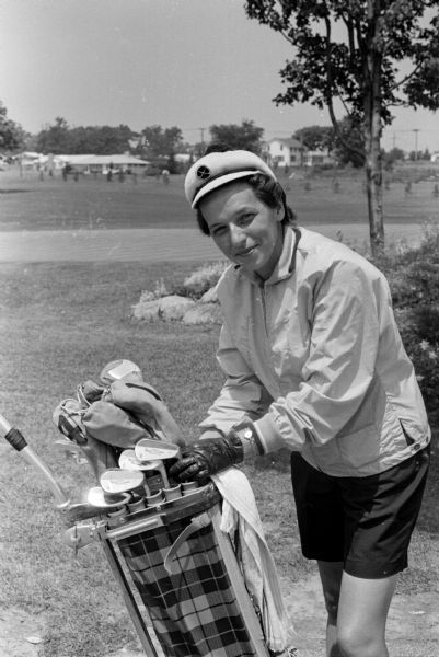 Shirley Tilsen models a sailor cap, golf jacket, and shorts while holding a plaid golf bag.