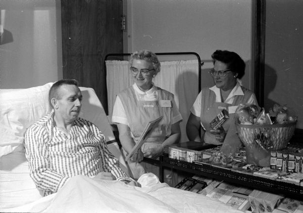 The Ladies in Pink Auxiliary operates a cart service for the patients Monday through Friday at St. Mary's Hospital. Mildred Downey, the incoming cart workers chairman, shows a magazine to George Vanderbleeman, a patient. Irene Coffee, right, is a buyer for the carts. The Auxiliary, a non-sectarian volunteer group, will hold a fall membership tea and hospital tour Wednesday in Sacred Heart Hall of the hospital.