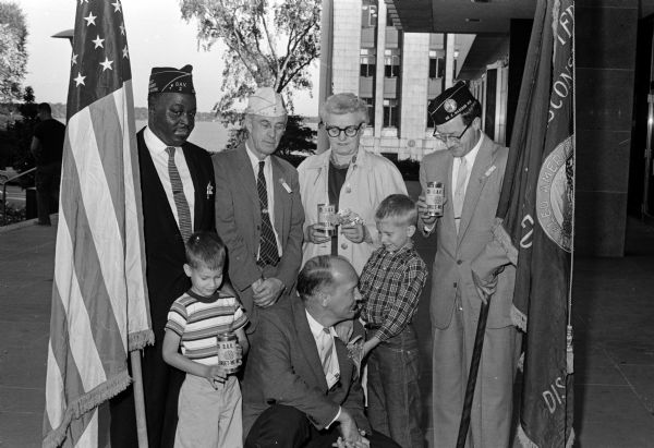 Madison Mayor Ivan A. Nestingen kneels next to two young boys, Steve and John Peckman, during a ceremony prior to the Forget-Me-Not fundraising drive of the Madison Chapter of the Disabled American Veterans. John presents the Mayor the first forget-me-not of the drive. In back are, left to right: John R. Parish, Earl Heath, Leone Wagner, and Howard Walstead.
