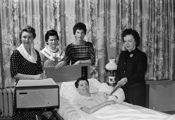 Pauline Dubner, a patient at the Wisconsin Neurological Foundation, poses in her hospital bed next to a new record player in a case. Behind her stand the four officers of the Madison Builders' Wives organization, who have gifted the foundation with a phonograph and five recorded books. The officers of the club are Margerat Dohm, Irene Daniels, Donna Smithback, and Muriel Jevne.