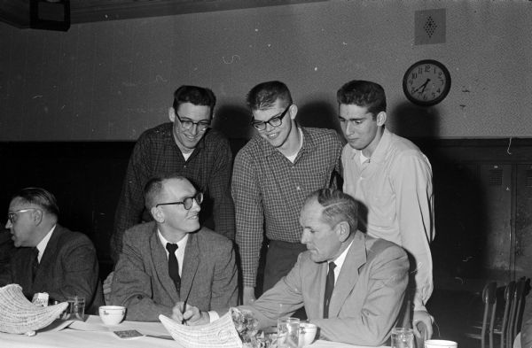 West High school's three star ends, standing, look over a football play drawn by Regent Coach Burt Hable, seated left, and Wisconsin assistant Coach and main speaker Clark Van Galder. The players, left to right, are: Ralph Farmer, Harry Kingsbury, and Henry Cuccia.