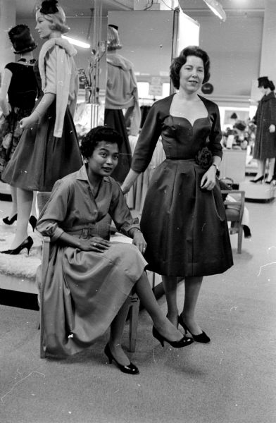 Two of the models who will display winter fashions at the YWCA style show and dessert party are Terry Alayu (seated), service director at Truax Field, and Ruth Smart. Terry Alayu is wearing a Dior blue silk shirtwaist dress and Ruth Smart is modeling a midnight blue peau de soie cocktail dress.