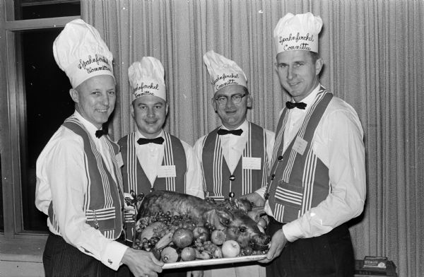 Four contractors are wearing chef's hats and aprons while showing off a roast suckling pig. They are, left to right: Dewey Badeau, Herman Hinrichs, Henry Johnson and Jerry Hill.  They are preparing for the Contractors 'Sphanferkel', a pork roast feast at which contractors meet socially with manufacturers, suppliers and sales representatives with whom they do business.   
