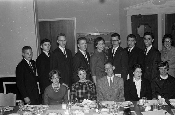 Group portrait of fourteen city and suburban youth honored by Madison's six Optimist Clubs at a banquet. Original caption states: "The fourteen selected represent the "95 per cent of America's young people who make major contributions to their communities each year." Standing, left to right ar:e Gary Johnson, Boy Scout drum and bugle corps; Gary Mahnke, Explorer Scout senior drum and bugle corps; Robert Swanson, Wisconsin High class president; Jack Porter, West High senate president. Mary Wright, West High student senate president; Dean Drury, Monona Grove student council president; John Voss, Edgewood student council president; David Reimer, East High youth council president; and Jeanne Weiler, Edgewood girls club president.  Seated are: Joan Wilkie, Wisconsin High student council and annual staff member; Betty Bakke, Central High girls club president; Ron Dintermann, Central High student council president; Karen Keeton, East High annual editor and Mary Vogt, Monona Grove student newspaper editor."