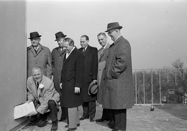 Secretary of State Robert Zimmerman, kneeling, presides at the cornerstone laying ceremony for the new Madison Inn, located at Frances and Langdon Streets. Six men look on, including from left to right: Al Steinhauer, president of Anchor Savings and Loan Assn.; Erwin Lackore, executive secretary of the Madison Chamber of Commerce; Frank Hausheer, vice-president of the State Chamber of Commerce; Conrad A. Elvejem, president of the University of Wisconsin; Ernest Lane, Milwaukee, president of the Madison Inn and Dorsey Botham, president of the Madison Chamber of Commerce.