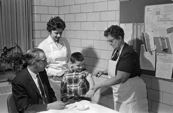 Dr. Joseph F. Behrend of Sun Prairie (left) and Carol Dorn, a public health nurse, look on as Mrs. R.J. McCarthy (right) administers a vaccine to Darrel Carpenter of Verona at an immunization clinic. Mrs. McCarthy is the chairman of the Dane County Association of Health Councils who sponsor volunteers that help the county public health nurses.