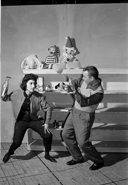 Sylvia Grunes raises a hammer threateningly towards O.J. Thompto during a performance of "The Puppet Prince." Grunes plays the Leprechaun and Thompto plays the evil magician, Magnus, who turns princes into puppets. Behind them are shelves of toys and puppets.