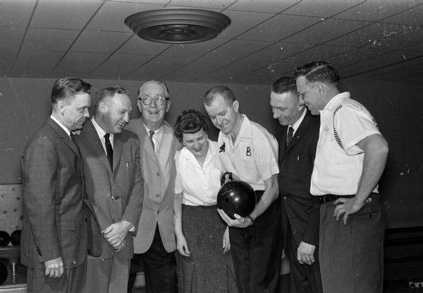 Group portrait of seven people at Stoughton's Badger Bowl bowling alley grand opening. At center wearing a bowling shirt is Harry Smith, 1960 national All-Star tourney champion, and a Brunswick adviser. Smith is holding a bowling ball and pointing out a detail in his bowling grip as Tootsie Erickson,leading Stoughton women bowler looks on. Also shown, left to right, are Olaf Roe, representing the city of Stoughton; Jim Grady, secretary of the Madison Bowling Assn.; Roundy Coughlin of the <i>Wisconsin State Journal</i>; and Dan Herbeck and Lyle Larson, co-managers  of the 'new 10-pin emporium.'