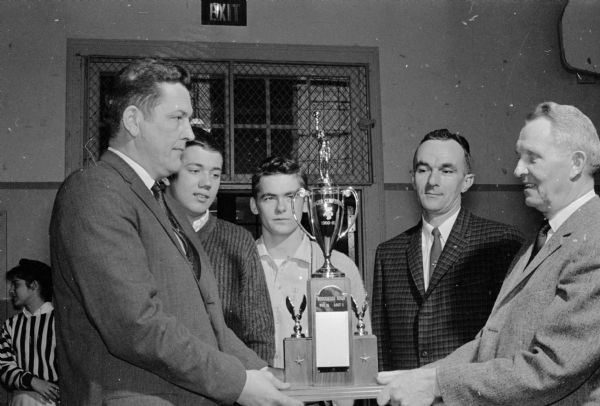 The Badger Conference basketball championship trophy is presented to Wisconsin High School. Shown (left to right) are: Warren Selbo, Stoughton High School principal and conference president; Pete Heebink, Wisconsin High most valuable player; Chuck Blake, team captain; Hal Metzen, coach; and John Goldgruber, principal.