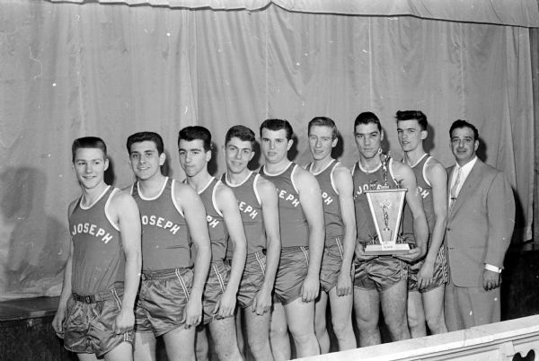 Indoor group portrait of the St. Joseph basketball team, winners of the championship of the Diocese of Madison Chi-Rho League. Players are, left to right: Fred Muehlemann,  Joe Provenzano, Jim Letourneau, John DiSalvo, Gary Schultz, Jim Deneen, Joe Engelhart, Dick Stoffels, and coach Ben Romano.