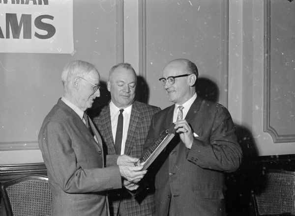 Three men examining a plaque. The original caption states: "F.J. Williams, left, Portage theater owner and longtime Wisconsin showman, receives a plaque commemorating his service to the Allied Theater Assn. of Wisconsin at a luncheon at Hotel Loraine. Williams, first president of the Wisconsin group, received the plaque from Edward Johnson, Milwaukee, right, national president of the group. S.J. Goldberg, center, a national director, is looking on. About 100 persons attended the spring meeting."