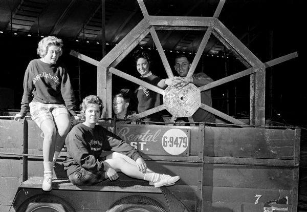 Five university students pose around a stage prop showboat paddle wheel while preparing for Campus Carnival. Their booth was built by Kappa Alpha Theta sorority and Kappa Sigma fraternity. The students are, left to right: Diana Mantor, Gretchen Toeken, Tony Reynolds, Muffy Wittmack, and Dick Anderson.