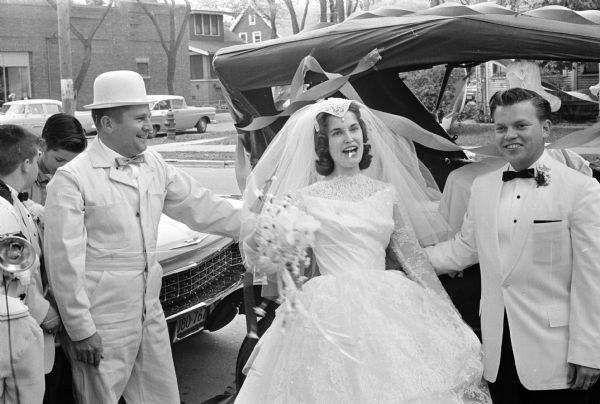 A newlywed couple in veil, wedding gown and white suit coat posing with the bride's uncle in front of a 1922 Model T Ford touring car. The couple, Mr. and Mrs. Dennis W. Day, were wed in St. Bernard's Catholic Church in Middleton. They were driven away by Benard Schroeder, who was wearing a 1920s "touring" costume.