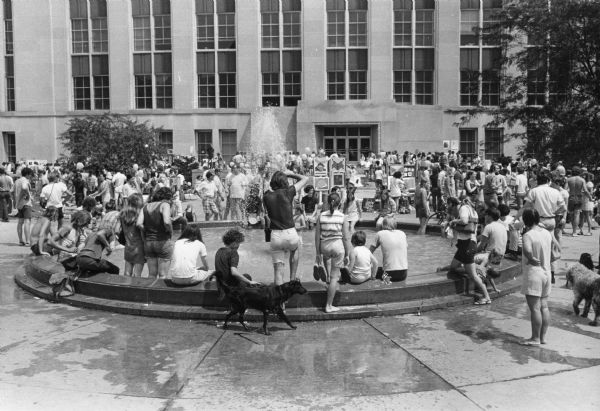 A crowd of people are cooling off in the Hagenah Fountain on Library Mall at the University of Wisconsin-Madison campus. The Memorial Library is in the background.
