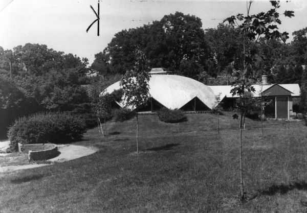 Exterior view of the Sunflower House at 5126 Tomahawk Trail, was designed and built by its first occupant, architect James Dresser in 1952. With a unique domed roof resembling a sunflower, the 40-foot diameter domed home is built of concrete over quonset ribs lined inside with redwood paneling. The lawn sloping towards the windows hides the sidewalls and affords added insulation. The dome is divided into three bedrooms, a bath, living room, and a 10-foot diameter kitchen below a central skylight.