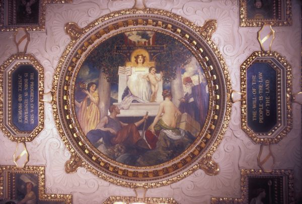 Painting by Hugo Ballin, called "Wisconsin Surrounded by her Attributes," on the Governor's Conference Room Ceiling at the Wisconsin State Capitol. The painting is an allegory of Wisconsin, depicted as a woman, with "Life, Liberty and the Pursuit of Happiness" written behind her head. She is surrounded by her attributes--beauty, strength, patriotism, labor, commerce, agriculture, and horticulture. The mottoes are "The Will of the People in the Law of the Land," "The Progress of a State is born in Temperance, Justice, and Prudence," and "Tempus Edax Rerum," (Time the devourer of all things).