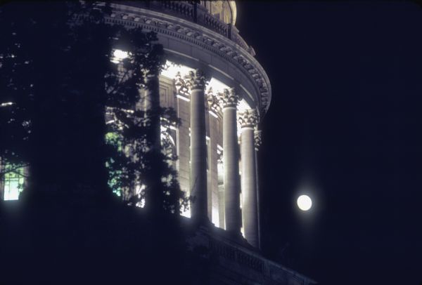 Colonnade around dome of the Wisconsin State Capitol at night with moon in background.