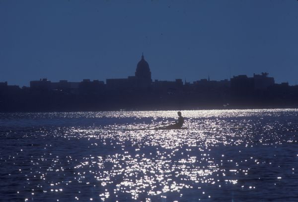 Man rowing scull in water in front of silhouette of the Madison skyline including the Wisconsin State Capitol.