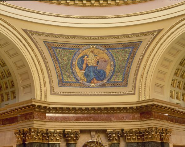"Legislation" pendentive, one of four panels of glass mosaic by Kenyon Cox, in the Wisconsin State Capitol. The artist sought the decorative properties of the Byzantine mosaics, while creating a style more in harmony with the purely Beaux-Arts character of the building. Located between the four arches in the rotunda, the pendentives make the transition from the octagonal form of the rotunda to the circular form of the dome. Each pendentive has a colossal seated figure on a gold background, which symbolize the three divisions of the powers of the state--the legislative, the executive, and the judicial. The last represents liberty, the foundation of all power in a free country.