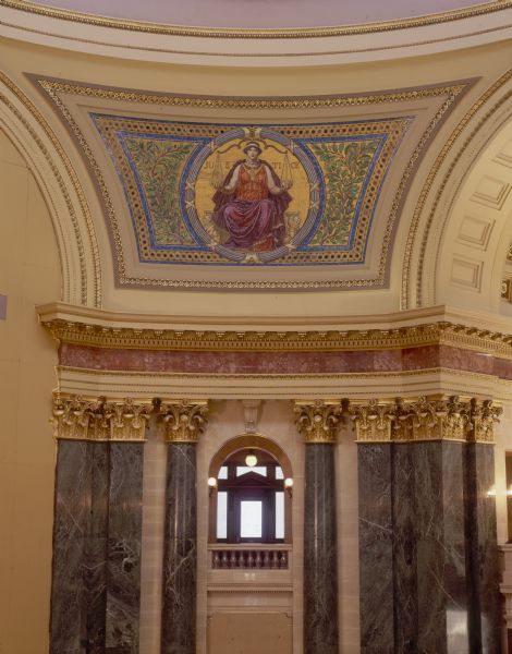 "Justice" pendentive, one of four panels of glass mosaic by Kenyon Cox, in the Wisconsin State Capitol. The artist sought the decorative properties of the Byzantine mosaics, while creating a style more in harmony with the purely Beaux-Arts character of the building. Located between the four arches in the rotunda, the pendentives make the transition from the octagonal form of the rotunda to the circular form of the dome. Each pendentive has a colossal seated figure on a gold background, which symbolize the three divisions of the powers of the state--the legislative, the executive, and the judicial. The last represents liberty, the foundation of all power in a free country.