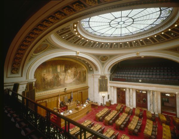 The Assembly Chamber from the visitors' gallery, in the Wisconsin State Capitol. There is a large circular ceiling skylight, and a mural painting by Edwin Howland Blashfield, representing Wisconsin.