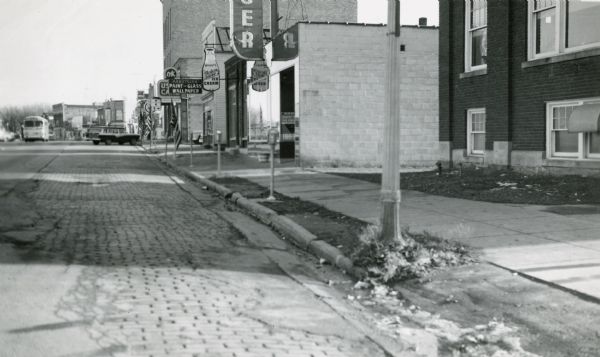 Photograph taken by the State Highway Department to show the condition of curbs and gutters along STH 12 in Mauston, also documenting the original brick surface and an unusual array of signs.