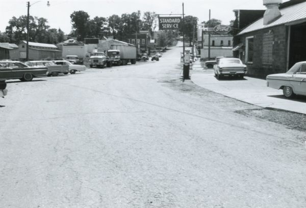 Monroe County Highway P in Kendall, Wisconsin, looking south.  The view includes an automobile service station and dealership, with automobiles and trucks randomly parked across the street, apparently waiting for repairs.