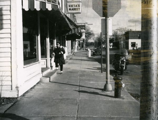 View of a woman walking on the sidewall along Bridge Street in New Lisbon.  The woman is about to enter a shop with an old storefront.  Behind her is a sign advertising Kieta's Market.