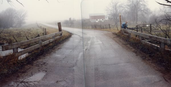 Elm Valley Road Bridge over a branch of the Little Wolf River.  In the distance, a red barn can be seen through the fog. In the middle distance a member of the crew taking the photograph is inspecting part of the bridge.