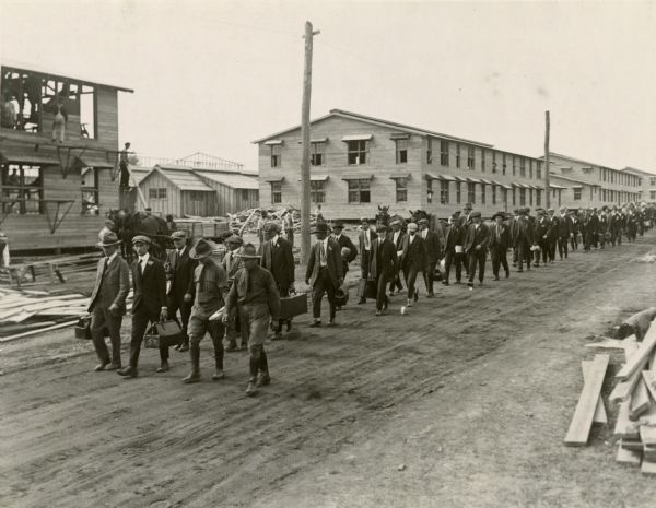 New army recruits still in their civilian clothes arriving at an unidentified World War I training camp. Many barracks, some still under construction, are visible in the background.
