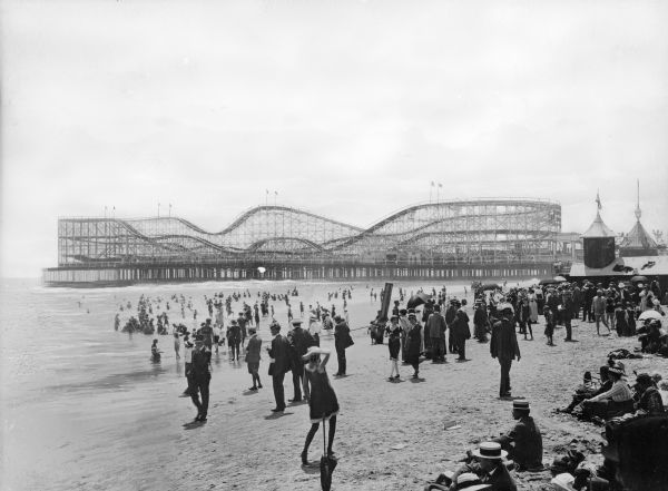 View of a crowd on the beach. The amusement park "The Pike" can be seen in the background, which was home to The Jack Rabbit Racer, which at the time was the world's second largest roller coaster.